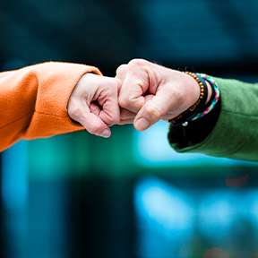 Two people perform a fist bump, one in orange and the other in green, against a blurred blue and green background.
