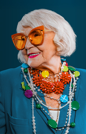 Woman in her 70s, smiling, wearing colorful jewelry and sunglasses.