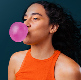 Person in an orange tank top blows a pink bubblegum bubble against a dark teal background.