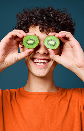 Person with curly hair holds kiwi halves over eyes, smiling, wearing an orange shirt against a blue background.