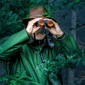 Man in green with hat uses binoculars in a dense, lush forest setting.