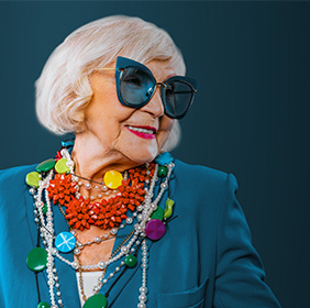 Woman in her 70s, smiling, wearing colorful jewelry and sunglasses.
