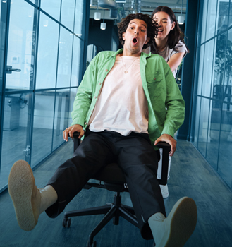 Woman pushing young man, with a surprised look on his face, through a hallway on an office chair.