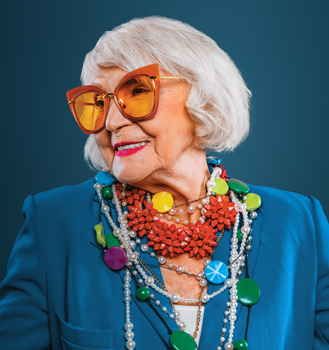 Woman in her 70s, smiling, wearing colorful jewelry and sunglasses.